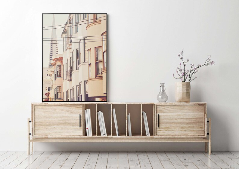 May include: A light wood cabinet with two sliding doors and a center shelf with books. A framed photograph of a city street with buildings and power lines hangs above the cabinet. A vase with a small bouquet of purple flowers sits on the cabinet.