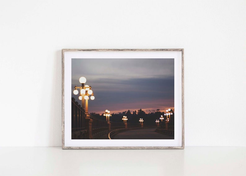 May include: A photograph of a street lined with lampposts at dusk. The lampposts are lit, casting a warm glow on the street. The sky is a soft purple and pink, with clouds in the distance.