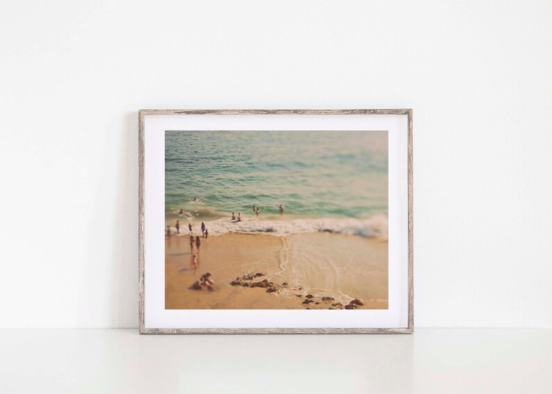 May include: A framed photograph of a beach scene with people swimming in the ocean. The photo is in a light wood frame and has a vintage feel.