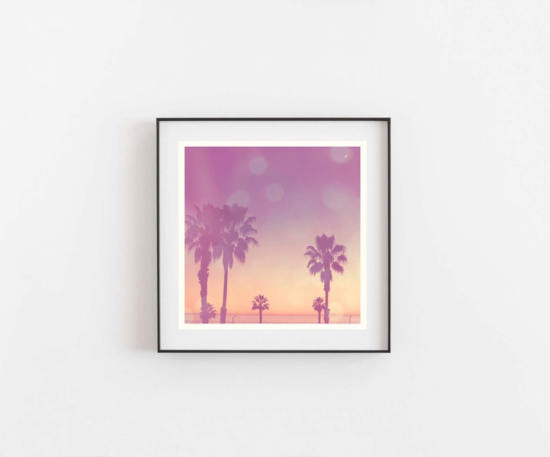 Op de afbeelding: Ingelijste print met een tropische zonsondergang. Palmbomen silhouetten tegen een lucht van roze, paarse en oranje tinten. Het kunstwerk roept een gevoel van rust en kustschoonheid op, perfect voor interieurdecoratie.
