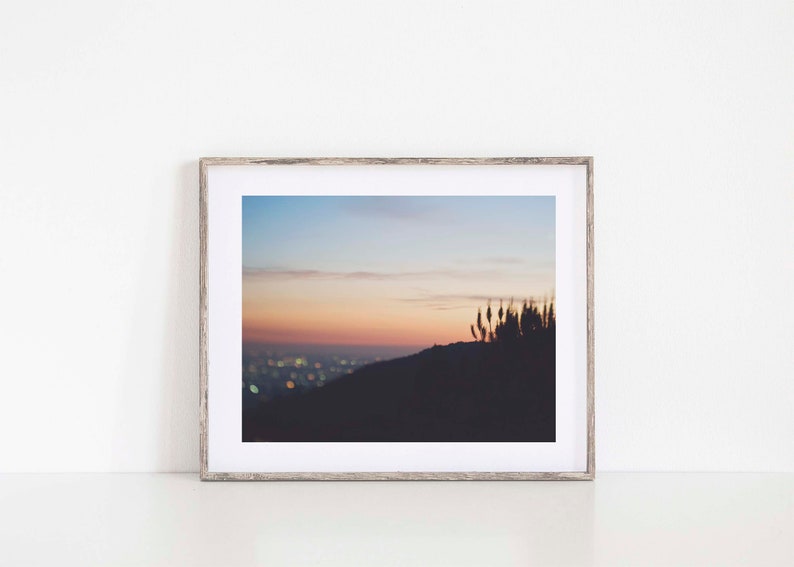 Op de afbeelding: Een ingelijste foto van een skyline bij zonsondergang. De stadslichten zijn wazig in de verte en de lucht is zacht roze en oranje. Silhouetten van hoog gras zijn op de voorgrond te zien.