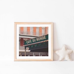 May include: A framed print featuring the Cafe Du Monde sign in New Orleans. The sign is green with white lettering and small lights. The background shows a brick building with balconies. A star-shaped pillow rests on the white surface.