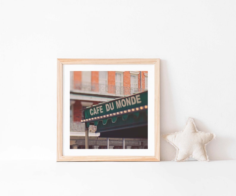 May include: A framed print featuring the Cafe Du Monde sign in New Orleans. The sign is green with white lettering and small lights. The background shows a brick building with balconies. A star-shaped pillow rests on the white surface.