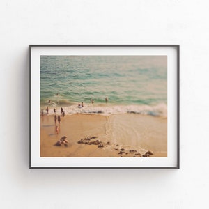 May include: A framed photograph of a beach scene with people swimming and sunbathing in the ocean. The photo is taken from a high vantage point, looking down at the beach. The water is a light blue color, and the sand is a light brown color. The sky is a light blue color.