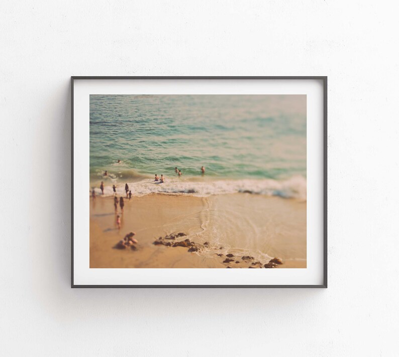 May include: A framed photograph of a beach scene with people swimming and sunbathing in the ocean. The photo is taken from a high vantage point, looking down at the beach. The water is a light blue color, and the sand is a light brown color. The sky is a light blue color.