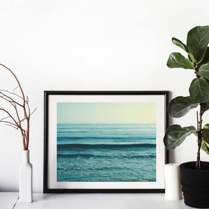 May include: A black framed photograph of a blue ocean with waves crashing on the shore. The photo is hanging on a white wall with a plant and a vase in the foreground.