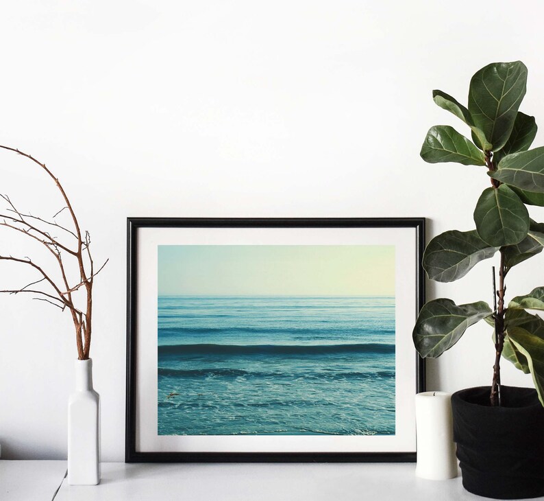 May include: A black framed photograph of a blue ocean with waves crashing on the shore. The photo is hanging on a white wall with a plant and a vase in the foreground.