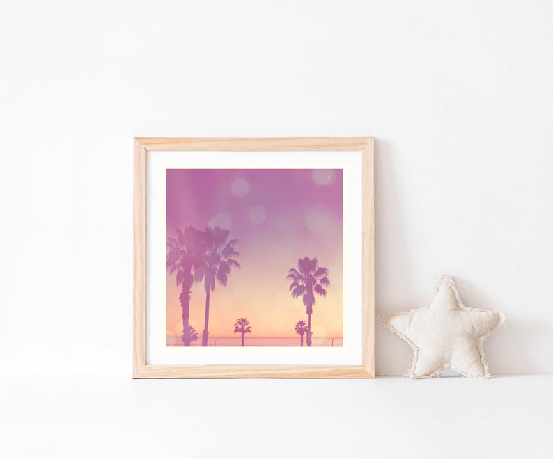 Op de afbeelding: Vierkantige ingelijste print met een zonsondergang met silhouetten van palmbomen. De lucht gaat over van paars naar oranje, met bokeh-lichten. Een beige stervormig kussen ligt rechts.