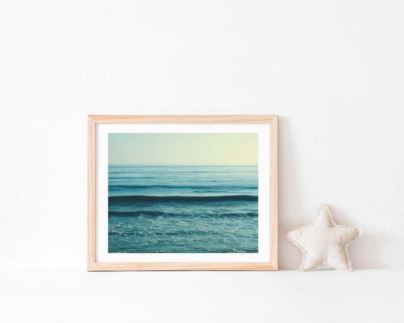 May include: A framed photograph of a blue ocean with waves crashing on the shore. The photo is in a light wood frame and is hanging on a white wall.