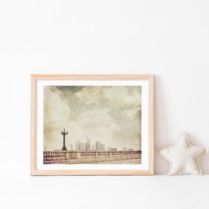 May include: A framed photograph of a cityscape with a vintage aesthetic. The image features a bridge with a street lamp and a view of a city skyline with buildings in the distance. The sky is cloudy and the overall tone is muted and nostalgic.