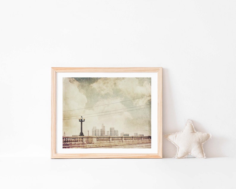 May include: A framed photograph of a cityscape with a vintage aesthetic. The image features a bridge with a street lamp and a view of a city skyline with buildings in the distance. The sky is cloudy and the overall tone is muted and nostalgic.