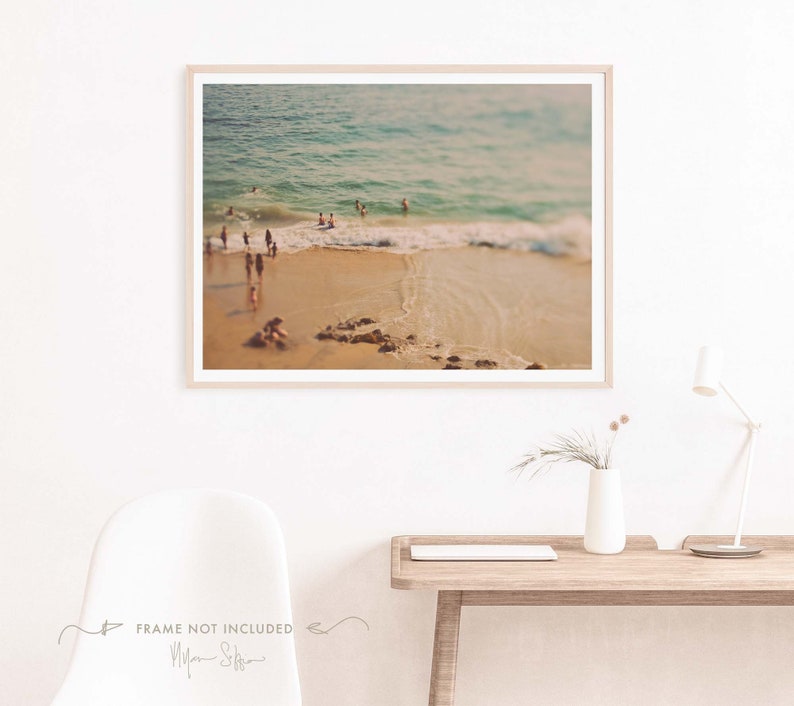 May include: A blurry photograph of a beach scene with people swimming in the ocean. The water is a light blue color and the sand is a light brown color. The photo has a vintage filter applied to it.