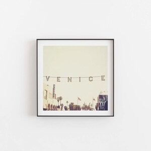 May include: A framed photograph of a street scene in Venice, California. The word "VENICE" is spelled out in large letters strung across the street. Palm trees line the street, and there are buildings in the background.