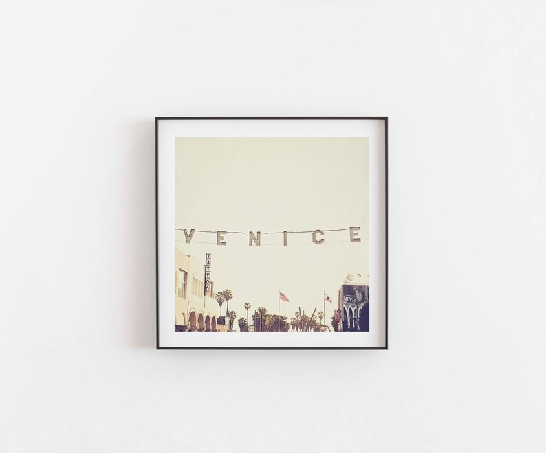 May include: A framed photograph of a street scene in Venice, California. The word "VENICE" is spelled out in large letters strung across the street. Palm trees line the street, and there are buildings in the background.