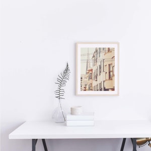 May include: A framed photograph of a city street with buildings in a warm color palette. The buildings have windows and are lined up in a row. There are power lines running across the top of the image.