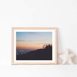 Op de afbeelding: Een ingelijste foto van een skyline bij zonsondergang. De stadslichten zijn in de verte zichtbaar en de lucht is zacht roze en oranje. De foto is ingelijst in een licht houten lijst.