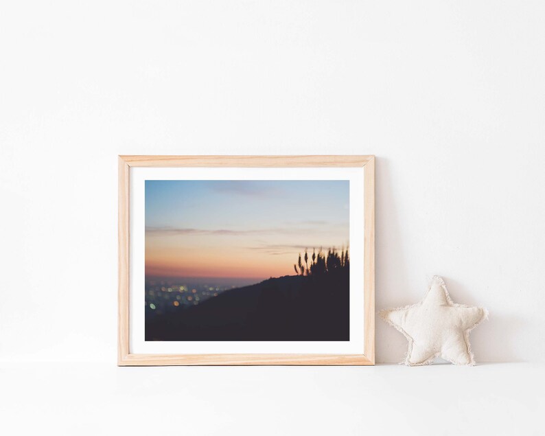 Op de afbeelding: Een ingelijste foto van een skyline bij zonsondergang. De stadslichten zijn in de verte zichtbaar en de lucht is zacht roze en oranje. De foto is ingelijst in een licht houten lijst.