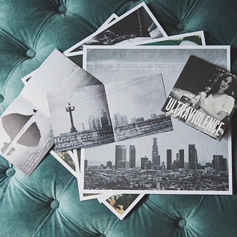 May include: A black and white photo of a city skyline with the text "Ultraviolence" printed on a black background. The photo is part of a stack of photos and papers, including a black and white photo of a street scene with a streetlight and a building in the background.