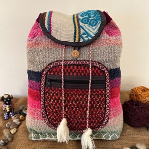 May include: A woven backpack featuring horizontal stripes in shades of pink, grey, and purple. The flap has a blue and white geometric pattern, and the front pocket is a dark red textured fabric with a zipper, two braided drawstrings, and white tassels.