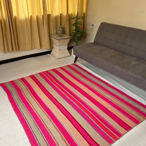 Puede incluir: Una alfombra rayada rosa y gris con textura tejida. La alfombra está colocada sobre un suelo blanco en una habitación con un sofá y una mesa auxiliar.