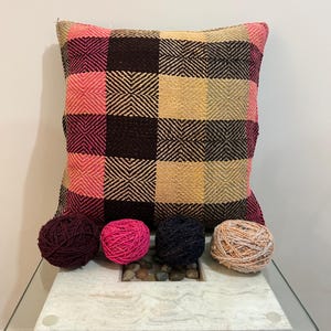 Puede incluir: Un cojín decorativo con un diseño a cuadros en tonos rosa, marrón y beige. Cuatro ovillos de lana en varios colores, incluyendo burdeos, rosa intenso y negro, están colocados frente al cojín, sugiriendo un tema artesanal.