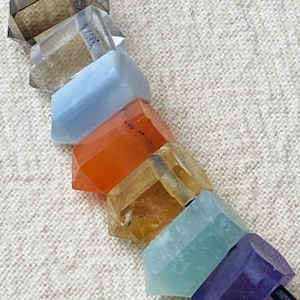 Könnte beinhalten: Eine Halskette mit sieben facettierten Edelsteinperlen in einer Regenbogenfarbenfolge. Die Perlen sind auf einer schwarzen Schnur aufgefädelt. Die Perlen sind: klar, hellbraun, hellblau, orange, gelb, hellgrün und lila.