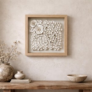 May include: A square, framed wall art piece featuring a collection of white, textured flowers. Two large rose-like blooms are positioned in the upper left corner, with smaller flowers filling the rest of the frame. The frame is made of light-colored wood.