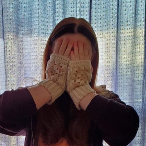 Puede incluir: Una persona lleva un par de guantes sin dedos de ganchillo de color crema. Los guantes tienen un patrón cuadrado y cubren las manos de la persona.