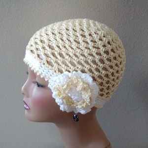 May include: A crocheted hat in a pale yellow color with a white crocheted flower on the side. The hat is made with a mesh stitch and has a simple design.