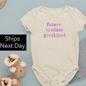 May include: A cream-colored baby onesie with the text "future madam president" in pink, blue, and orange.