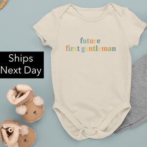 May include: A cream-colored baby onesie with the text "future first gentleman" in rainbow colors.