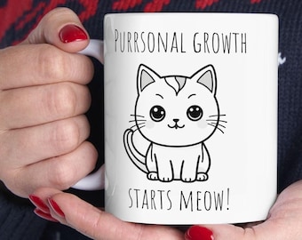 May include: White ceramic mug with the text "PURRSONAL GROWTH STARTS MEOW!" and a cartoon cat illustration. The cat is black and white with large eyes and a smiling face. The mug is held by a person.