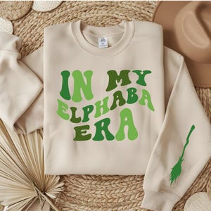 May include: A beige sweatshirt with the text "In My Elphaba Era" in green letters. The letters are in a retro, wavy font. A green broom is printed on the right sleeve.