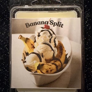 May include: A wax melt in a clear plastic clamshell package. The label features a banana split dessert with ice cream, bananas, and a cherry. The words "Banana Split" are printed above the image.