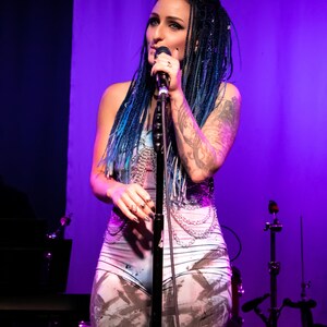 May include: A performer with blue-streaked hair sings into a microphone on a stage with purple lighting. She wears a light-colored bodysuit with a chain design and a tattoo on her arm. The stage has musical equipment.