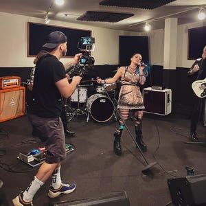 May include: A band performing in a recording studio. The lead singer, with blue hair, is holding a microphone. A person is filming the band with a camera. Other band members are playing instruments, including a guitar and drums.