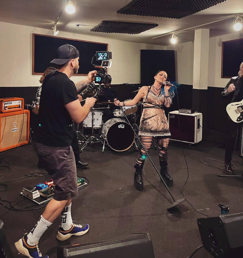 May include: A band performing in a recording studio. The lead singer, with blue hair, is holding a microphone. A person is filming the band with a camera. Other band members are playing instruments, including a guitar and drums.