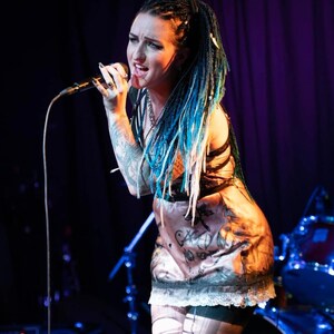 May include: A woman with blue-streaked braids sings into a microphone on stage. She wears a patterned dress with a lace hem and patterned stockings. Her arms are covered in tattoos. The background is dark with stage lighting.
