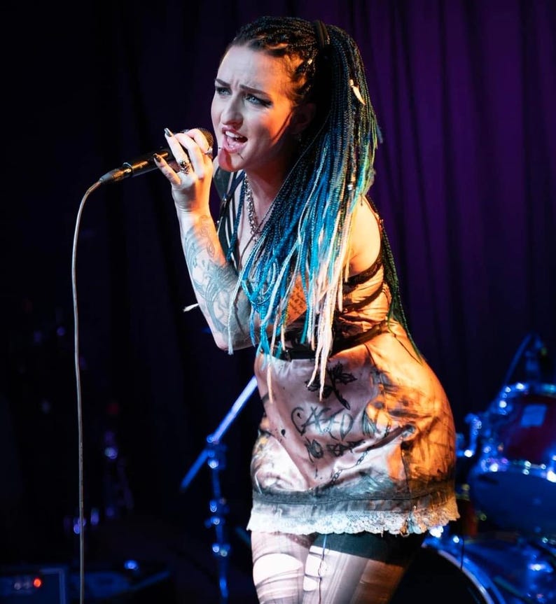 May include: A woman with blue-streaked braids sings into a microphone on stage. She wears a patterned dress with a lace hem and patterned stockings. Her arms are covered in tattoos. The background is dark with stage lighting.
