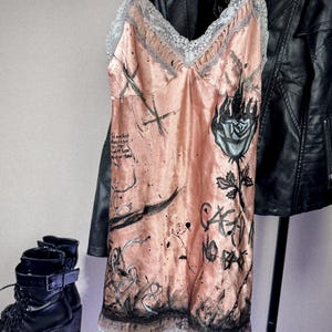 May include: A peach-colored slip dress with a lace trim and spaghetti straps. The dress features black graffiti-style artwork, including a rose and text. A black leather jacket hangs in the background.