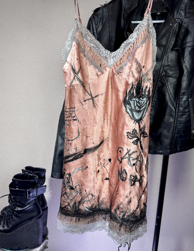 May include: A peach-colored slip dress with a lace trim and spaghetti straps. The dress features black graffiti-style artwork, including a rose and text. A black leather jacket hangs in the background.