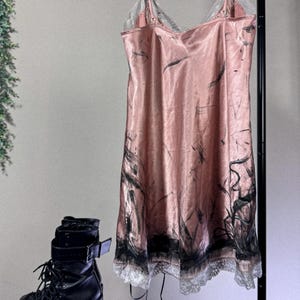 May include: A pink satin slip dress with black abstract brushstrokes and lace trim hangs on a hanger. Black platform boots with laces and buckles are in the foreground. The dress has spaghetti straps.
