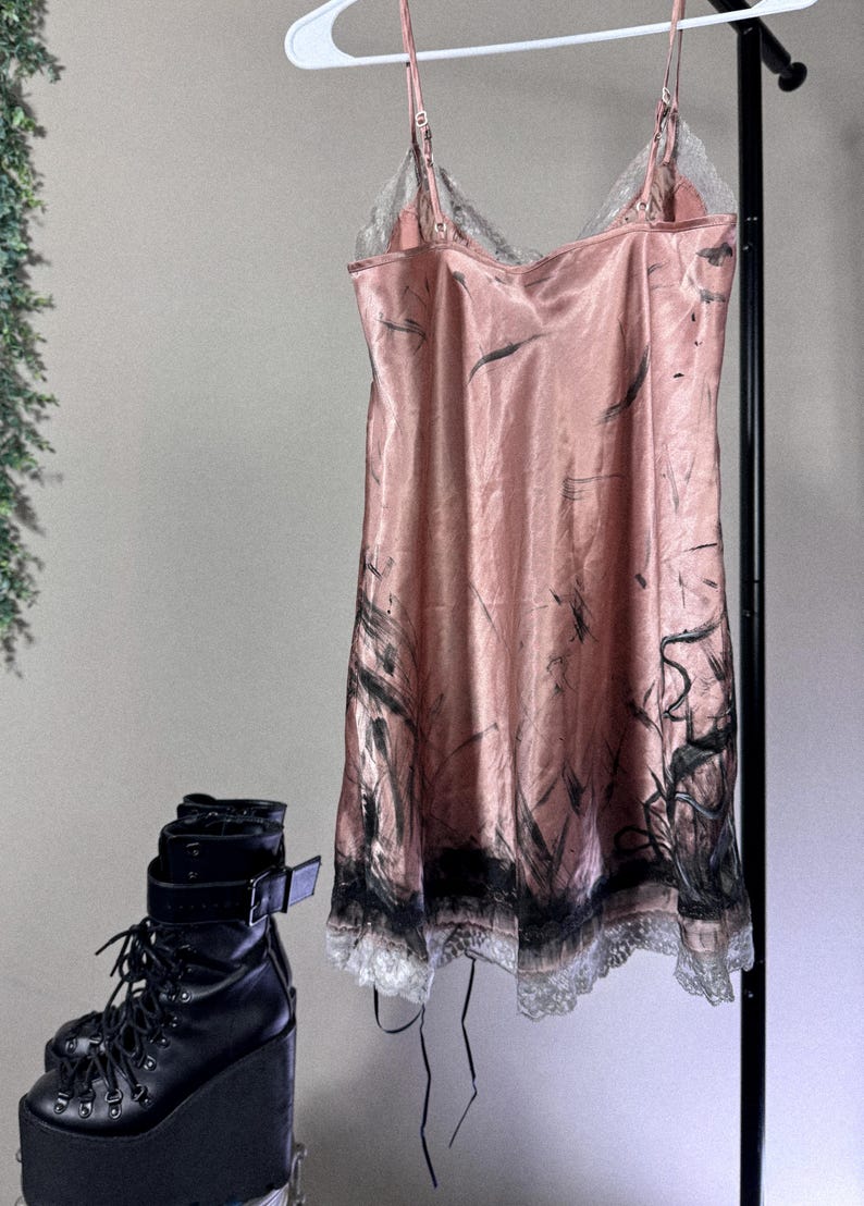 May include: A pink satin slip dress with black abstract brushstrokes and lace trim hangs on a hanger. Black platform boots with laces and buckles are in the foreground. The dress has spaghetti straps.