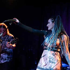 May include: A woman with blue and green dreadlocks and tattoos performs on stage with a microphone. She wears a pink dress with the words "CARD IN NO BARS" and a black top. A man plays a guitar in the background.