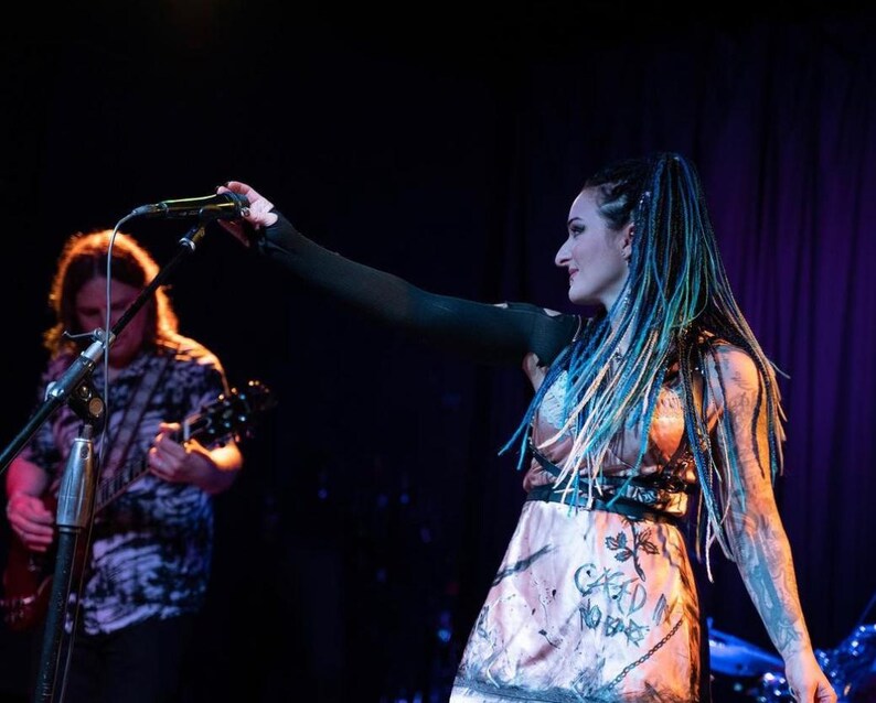 May include: A woman with blue and green dreadlocks and tattoos performs on stage with a microphone. She wears a pink dress with the words "CARD IN NO BARS" and a black top. A man plays a guitar in the background.