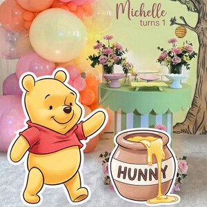 Puede incluir: Una escena de fiesta con el tema de Winnie the Pooh. Un recorte de Winnie the Pooh está en primer plano, con un tarro de miel que dice "HUNNY". Un arco de globos en tonos de rosa, naranja y amarillo está a la izquierda. Una mesa con flores y un telón de fondo con el texto "Michelle turns 1" está en el fondo.