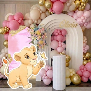 May include: A party backdrop featuring a cartoon lion cub, pink and gold balloons, and a white arch with the name "Olivia" in gold. The backdrop includes a pink panel with floral arrangements and a butterfly.