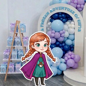 Puede incluir: Un recorte de dibujos animados de Anna de Frozen, con una capa morada y un vestido verde, se encuentra frente a un arco de globos. Una estantería de madera exhibe cajas de regalo con diseños de copos de nieve. El arco dice "A Frozen Adventure of Gifts."