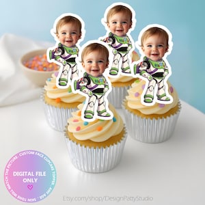 May include: Four cupcakes in silver foil liners topped with white frosting and colourful sprinkles. Each cupcake features a custom paper topper showing a baby's face superimposed onto a cartoon space ranger suit with green, purple, and white details.