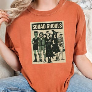 May include: Burnt orange t-shirt with a graphic of five stylized ghouls and the text "SQUAD GHOULS" in a rectangular frame. The ghouls include a zombie, vampire, witch, and skeleton. The shirt is made of a soft material.